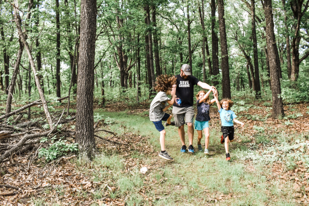 Three kids play with their dad & are laughing. One hasn't landed yet & his long hair is flying in the wind. The girl is laughing while holding her dad's hand. And the youngest little boy is laughing hysterically. The dad is looking down watching his kids.